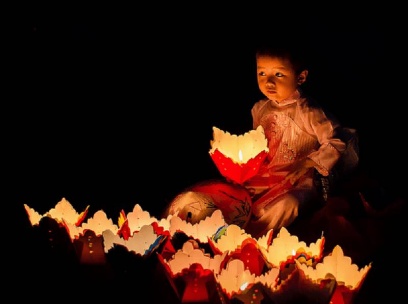 Những em bé nhỏ tuổi phụ cha mẹ sau giờ học ngồi gần mé sông bán đèn hoa đăng
